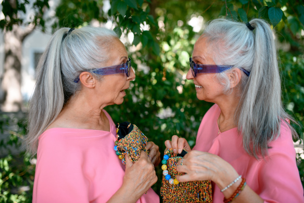 Senior women twins in colourful clothes with beads handbags in city, talking and smiling.