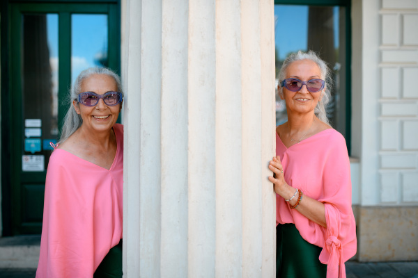 Senior women twins in colourful clothes in a city, posing next to architectural column.