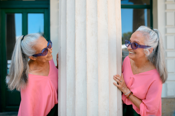 Senior women twins in colourful clothes in a city, posing next to architectural column.