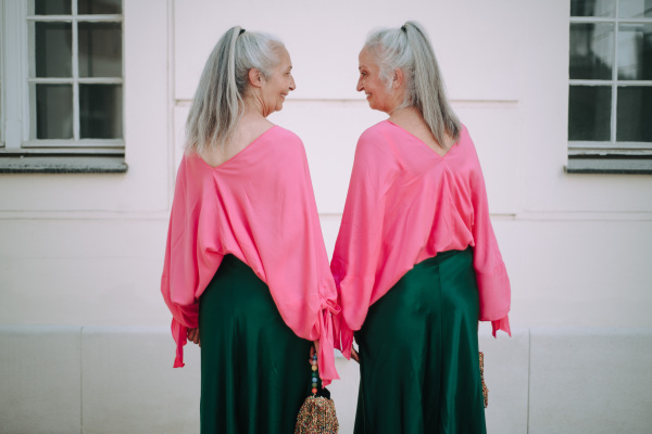 Senior women twins in colourful clothes in the city, shopping. Rear view.