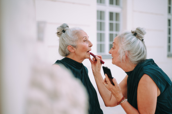 Senior women, twins in a city walk, completing make-up with lipstick.