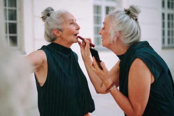 Senior women, twins in a city walk, completing make-up with lipstick.