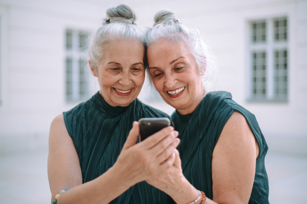 Senior women twins outdoors in city checking a smartphone.