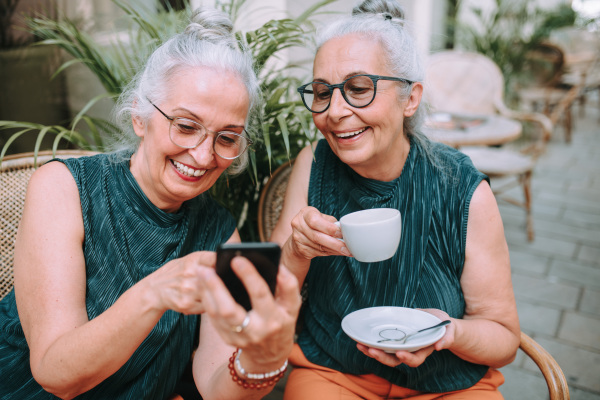 Happy senior women twins having coffee break in city, smiling and checking a phone.