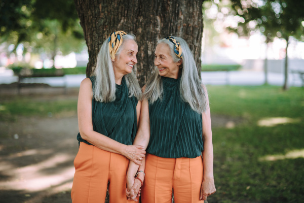 Happy seniors women, twins posing in front of a tree in city park, smiling and talking.