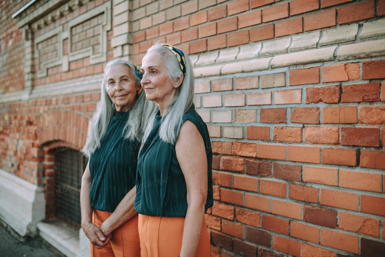 Senior women twins,in same clothes standing and posing in front of a brick wall, outdoor in city.