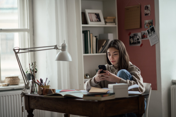 Young teenage girl doing homework and scrolling smartphone in her room.