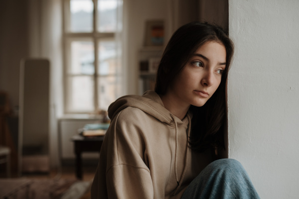 Unhappy teenage girl sitting in home and looking out of the window.