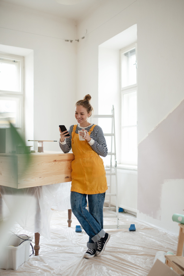 Happy young woman having break during renovation her house. Using phone, searching inspiration at social medias.