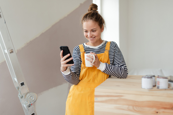 Happy young woman having break during renovation her house. Using phone, searching inspiration at social medias.