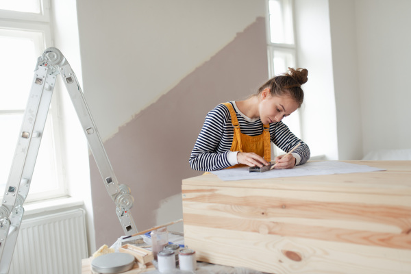 Happy young woman remaking wooden cabinet in her house. Concept of reusing materials and sustainable lifestyle.