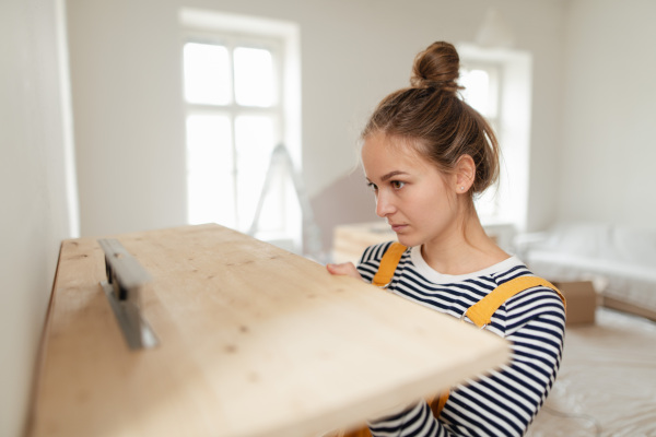 Young woman remaking her apartment, measuring wall with spirit level and hanging a shelf.