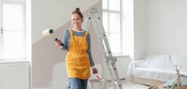 Happy young woman remaking her new house, painting walls. Concept of renovation, indepent women and sustainable lifestyle.