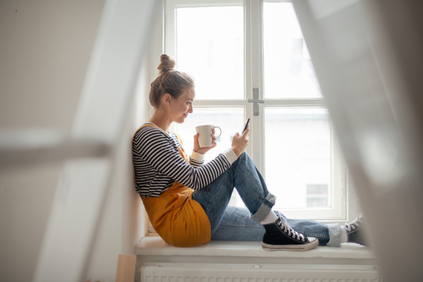 Happy young woman having break during renovation her house. Using phone, searching inspiration at social medias.