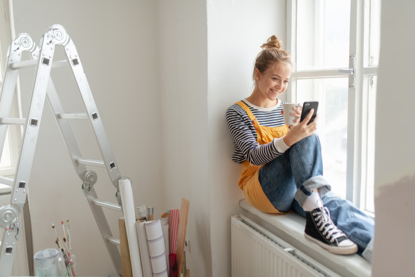 Happy young woman having break during renovation her house. Using phone, searching inspiration at social medias.