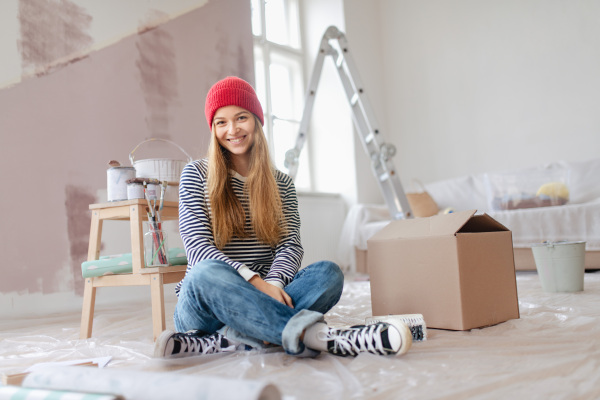 Happy young woman having break during painting walls in her house. Thinking about new projects. Concept of reusing materials and sustainable lifestyle.