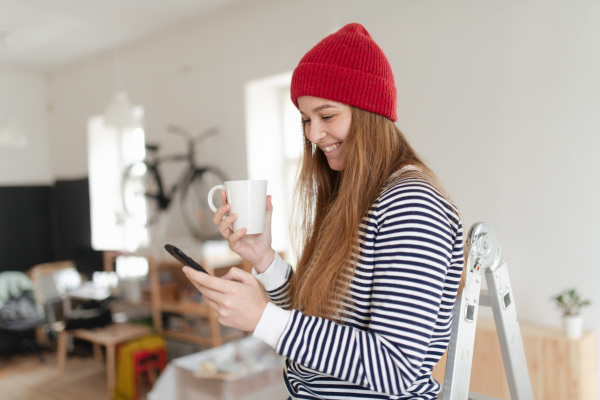 Happy young woman having break during renovation her house. Using phone, searching inspiration at social medias.