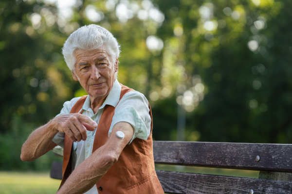 Senior man showing a sensor of continuous glucose monitor on his arm. Man with diabetes checking his blood glucose level outdoors, in city park.