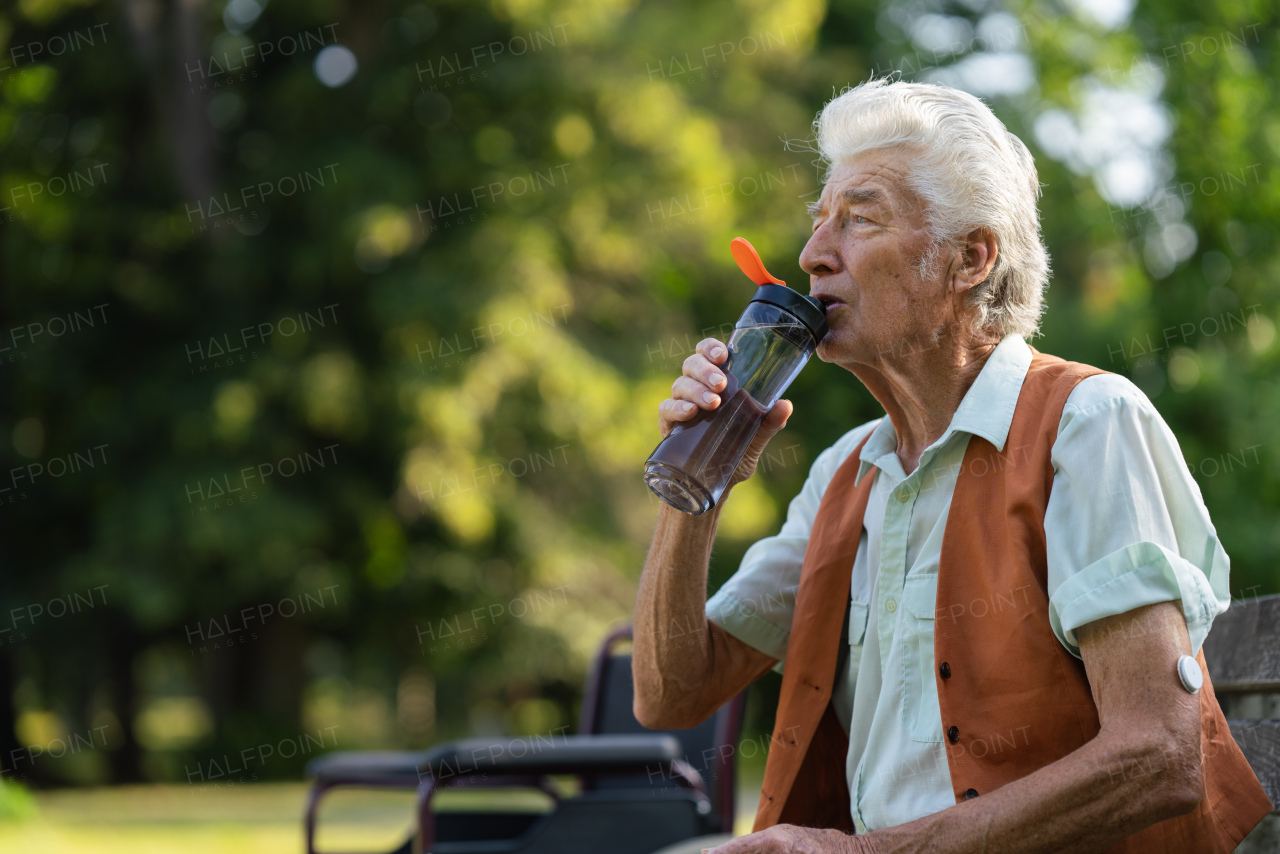 Diabetic senior patient using continuous glucose monitor to check blood sugar level at home. Senior man drinking water outdoor to better manage his diabetes. CGM device making life of elderly man easier.