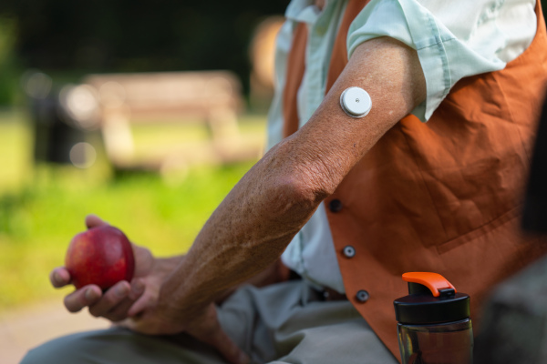 Close up of senior man with sensor of continuous glucose monitor on his arm. Man with diabetes checking his blood glucose level outdoors, in city park.