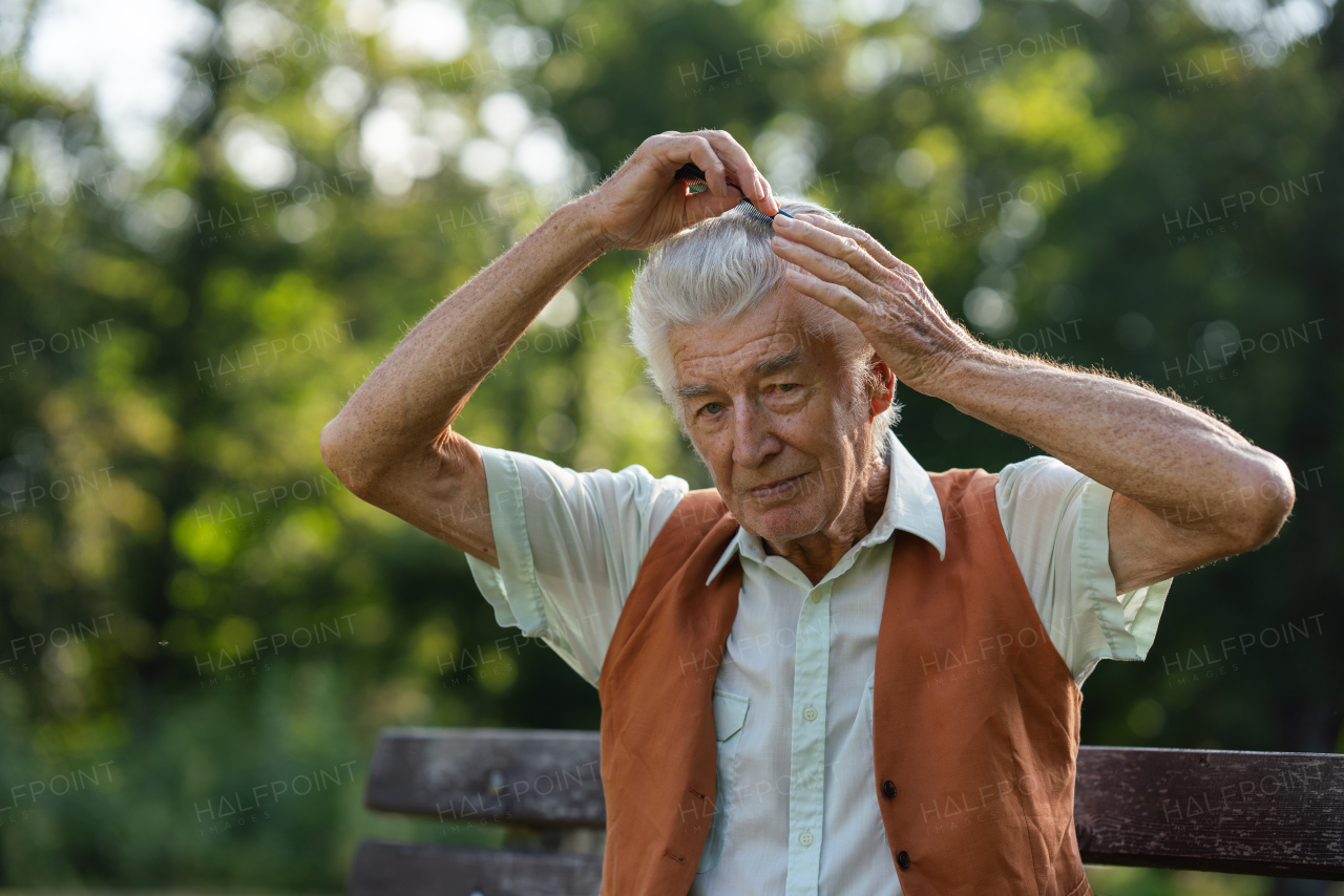 Senior man sitting on a bench in the park and combing his hair. Elderly man grooming himself before meeting someone. An older man with healthy, thick hair.