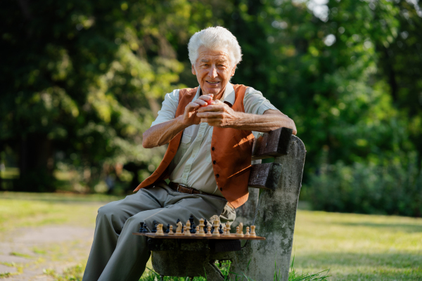 Portrait of senior man in a wheelchair is alone in a city park, feeling lonely. The man has a chessboard spread out on bench, playing by himself.