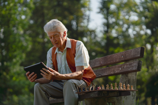 Senior man sitting in the park on a bench, looking at photos on his tablet and feeling nostalgic. The lonely elderly man waiting for a video call from his family.