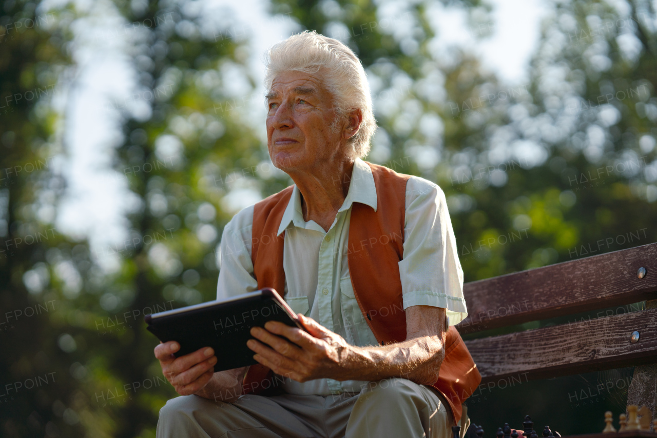 Senior man sitting in the park on a bench, looking at photos on his tablet and feeling nostalgic. The lonely elderly man waiting for a video call from his family.