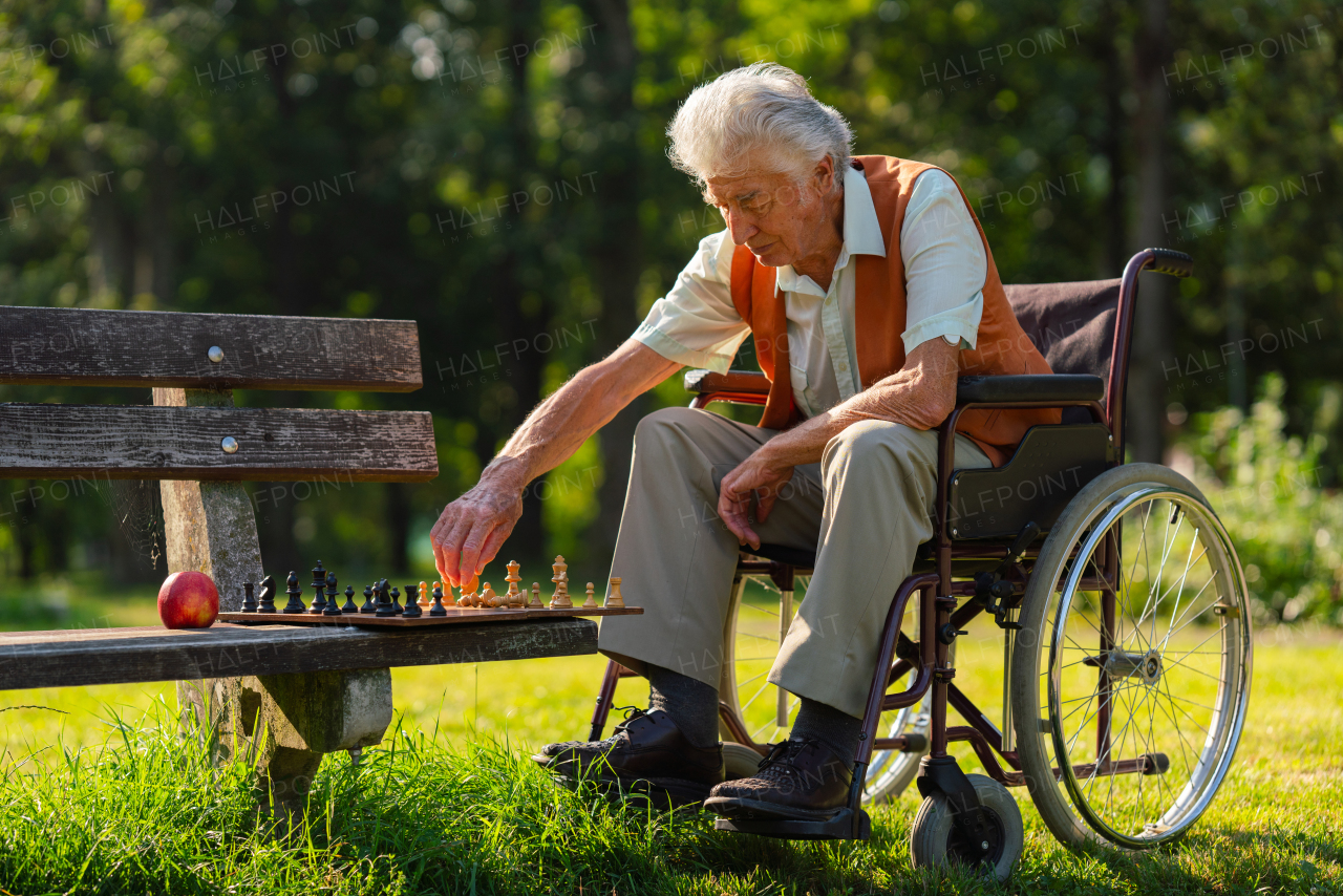 A senior man in a wheelchair is alone in a city park, feeling lonely. The man has a chessboard spread out on bench, playing by himself.