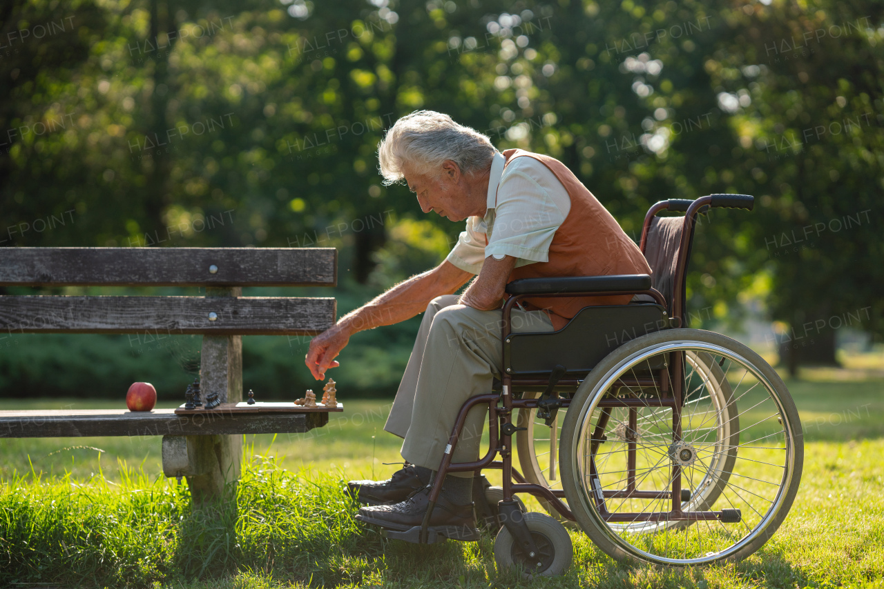 A senior man in a wheelchair is alone in a city park, feeling lonely. The man has a chessboard spread out on bench, playing by himself.