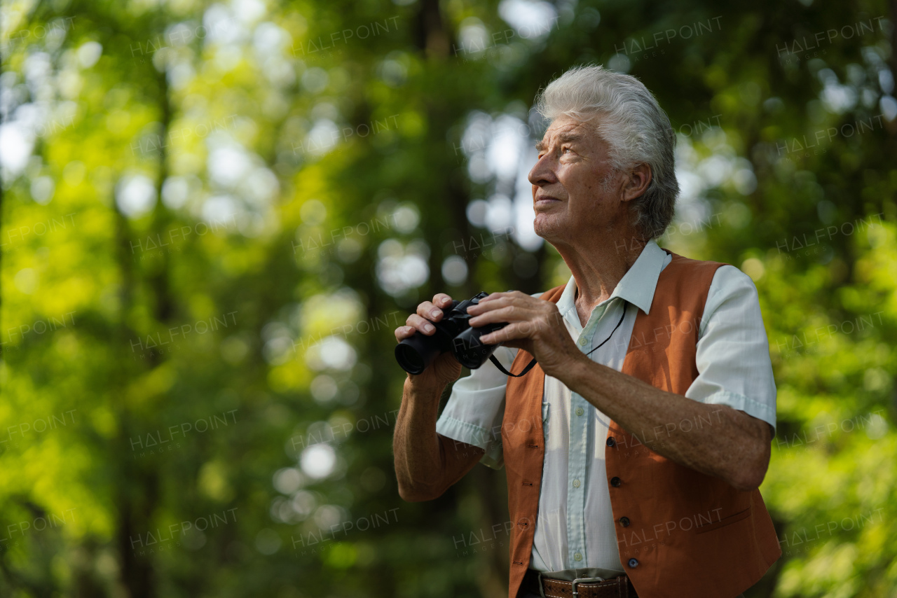 Senior man spending free time outdoors in nature, watching forest animals through binoculars. A retired forest ranger feeling at home in forest, monitoring wildlife and ecological changes.