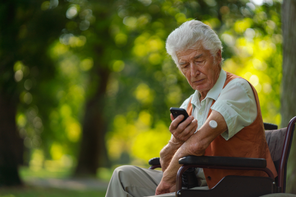 Senior diabetic man in wheelchair checking his glucose data on smartphone. Diabetic senior using continuous glucose monitor to check blood sugar level.