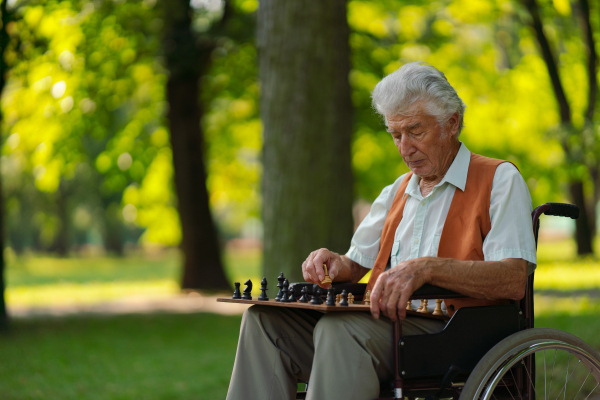 A senior man in a wheelchair is alone in a city park, feeling lonely. The man has a chessboard spread out on bench, playing by himself.