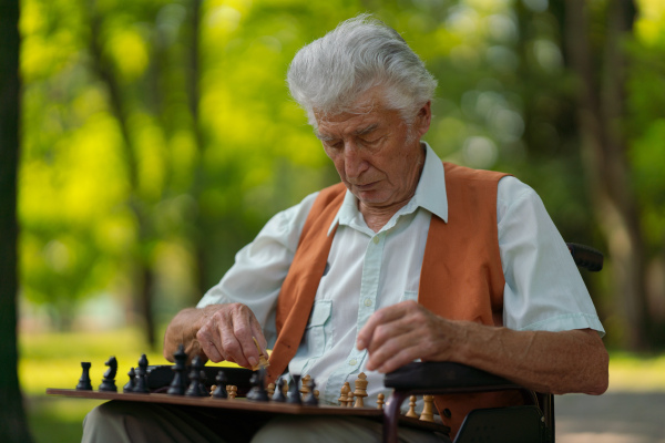 A senior man in a wheelchair is alone in a city park, feeling lonely. The man has a chessboard spread out on his lap, playing by himself.