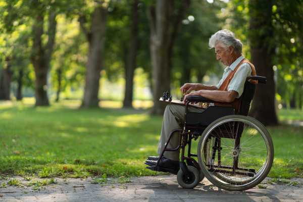 A senior man in a wheelchair is alone in a city park, feeling lonely. The man has a chessboard spread out on his lap, playing by himself.