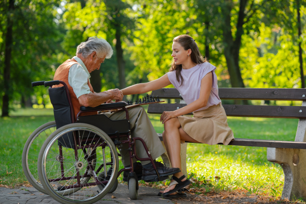 A young caregiver spending quality time with lonely senior client in the city park. The elderly man in a wheelchair spends time outdoors with his granddaughter, playing chess and chatting.