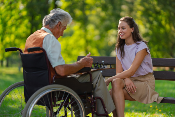 A young caregiver spending quality time with lonely senior client in the city park. The elderly man in a wheelchair spends time outdoors with his granddaughter, playing chess and chatting.