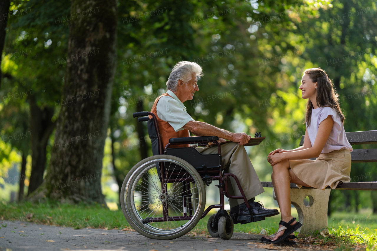 A young caregiver spending quality time with lonely senior client in the city park. The elderly man in a wheelchair spends time outdoors with his granddaughter, playing chess and chatting.