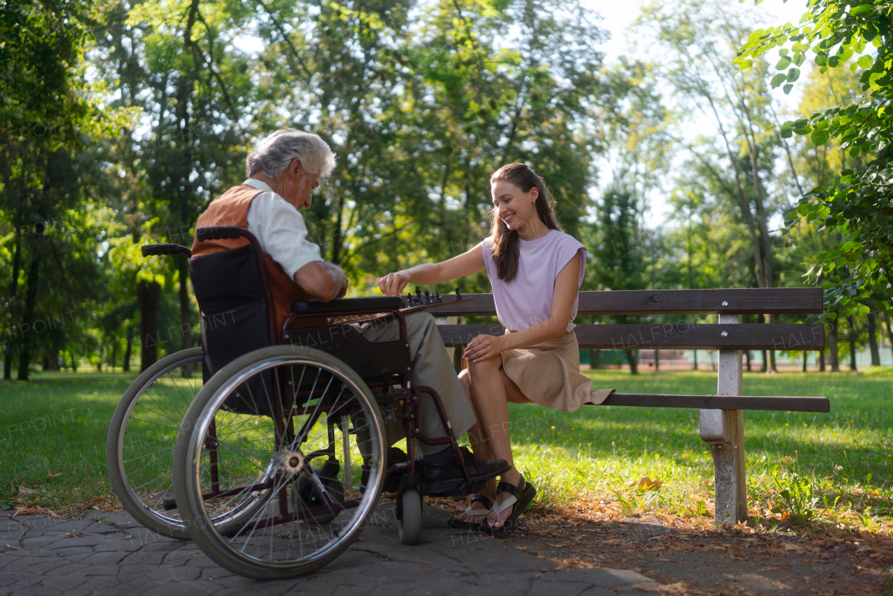 A young caregiver spending quality time with lonely senior client in the city park. The elderly man in a wheelchair spends time outdoors with his granddaughter, playing chess and chatting.