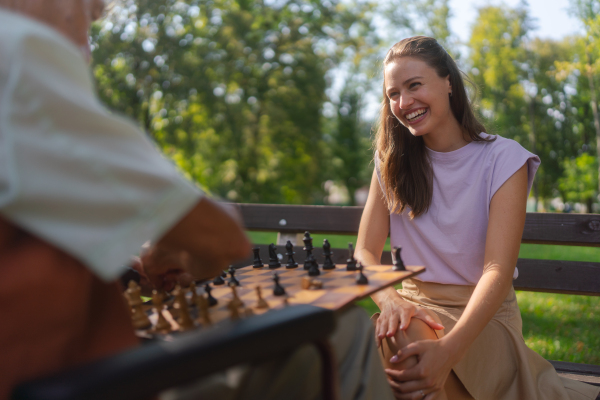 A young caregiver spending quality time with lonely senior client in the city park. The elderly man spending time outdoors with his granddaughter, playing chess and chatting.