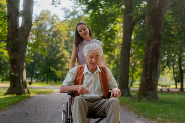 A young caregiver spending quality time with lonely senior client in the city park. Granddaughter pushing the grandfather in wheelchair in city park.