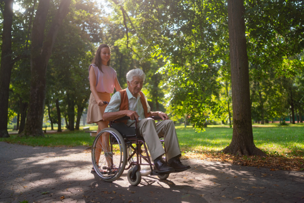 A young caregiver spending quality time with lonely senior client in the city park. Granddaughter pushing the grandfather in wheelchair in city park.