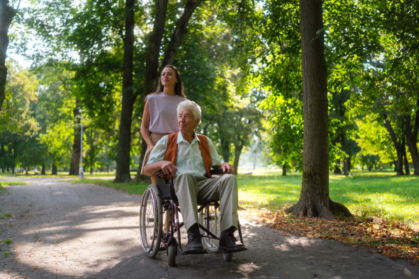 Granddaughter pushing the grandfather in wheelchair in city park. A young caregiver spending quality time with lonely senior client in the city park.