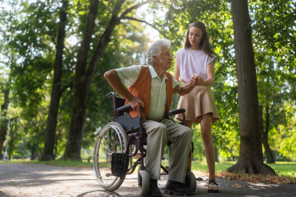 A young caregiver spending quality time with lonely senior client in the city park. Granddaughter helping the grandfather stand up from the wheelchair. The senior man is trying to stand and walk on his own, without the wheelchair.