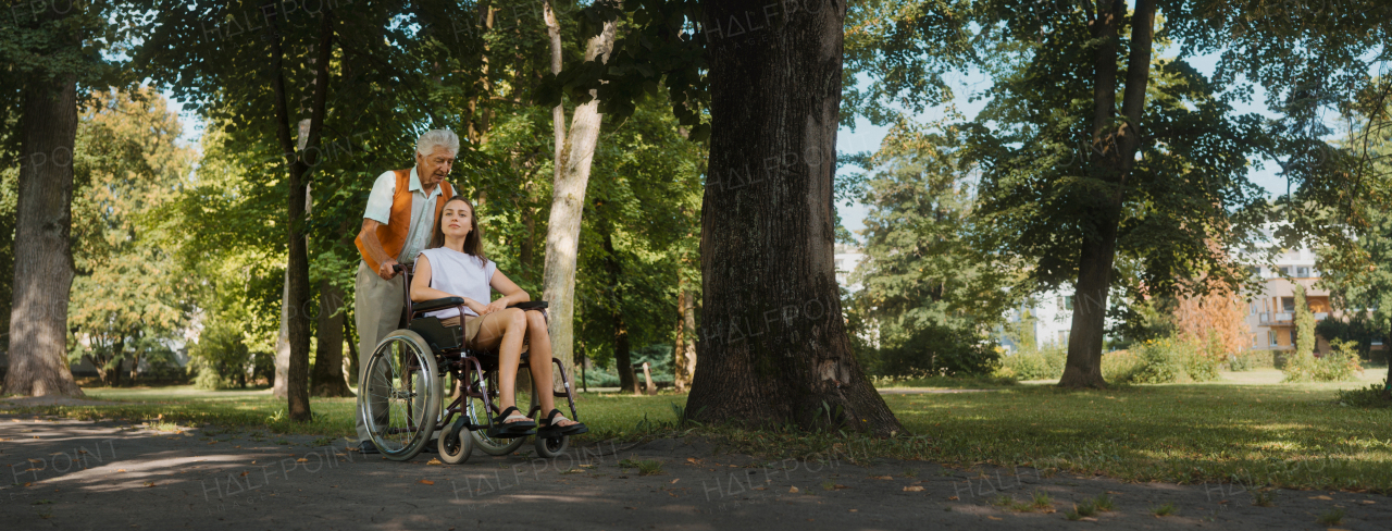 The senior man pushing young caregiver in a wheelchair. The elderly man feeling good and fit, he is able to push his granddaughter in the wheelchair. Banner with copy space.