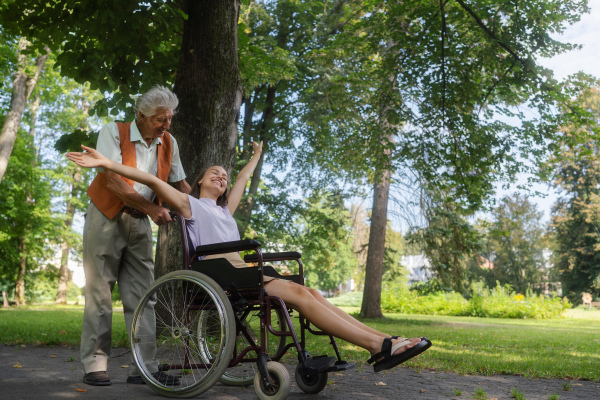 The senior man pushing young caregiver in a wheelchair, having fun. The elderly man feeling good and fit, he is able to push his granddaughter in the wheelchair.