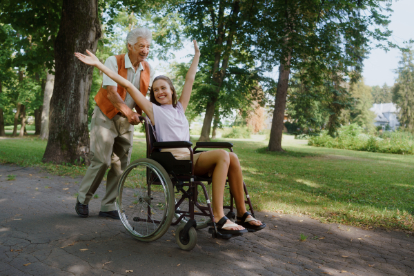 The senior man pushing young caregiver in a wheelchair, having fun. The elderly man feeling good and fit, he is able to push his granddaughter in the wheelchair.