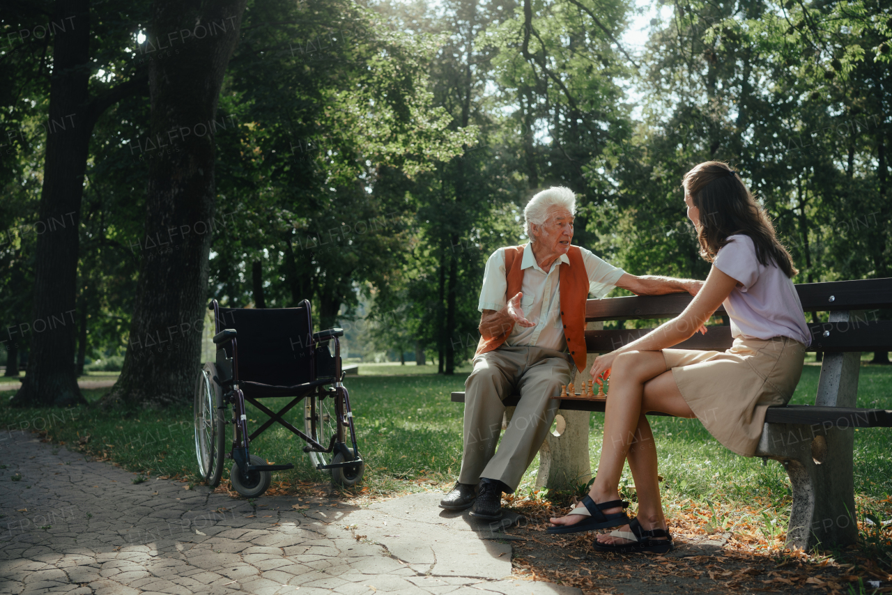 A young caregiver playing chess with lonely senior client in the city park. The elderly man in a wheelchair spends time outdoors with his granddaughter, playing chess and chatting.