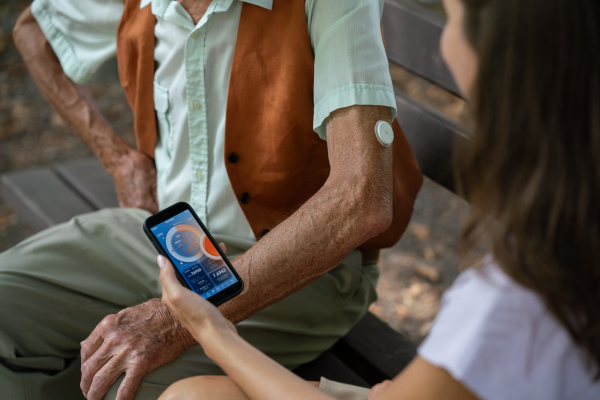 Caregiver helping senior diabetic man check his glucose data on smartphone. Diabetic senior using continuous glucose monitor. Granddaughter teaching her grandfather how to use smartphone, typing texts or browse.