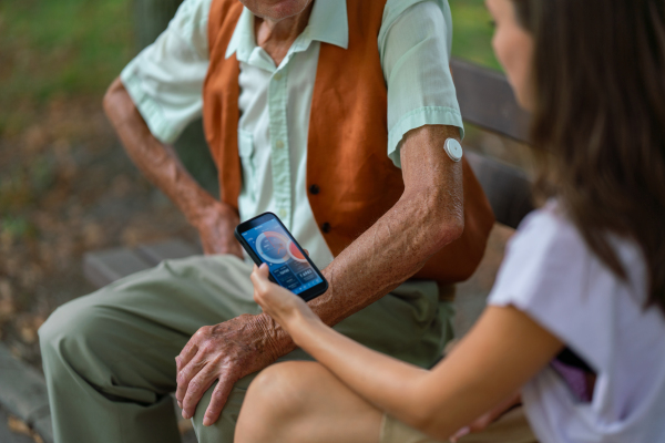 Caregiver helping senior diabetic man check his glucose data on smartphone. Diabetic senior using continuous glucose monitor. Granddaughter teaching her grandfather how to use smartphone, typing texts or browse.